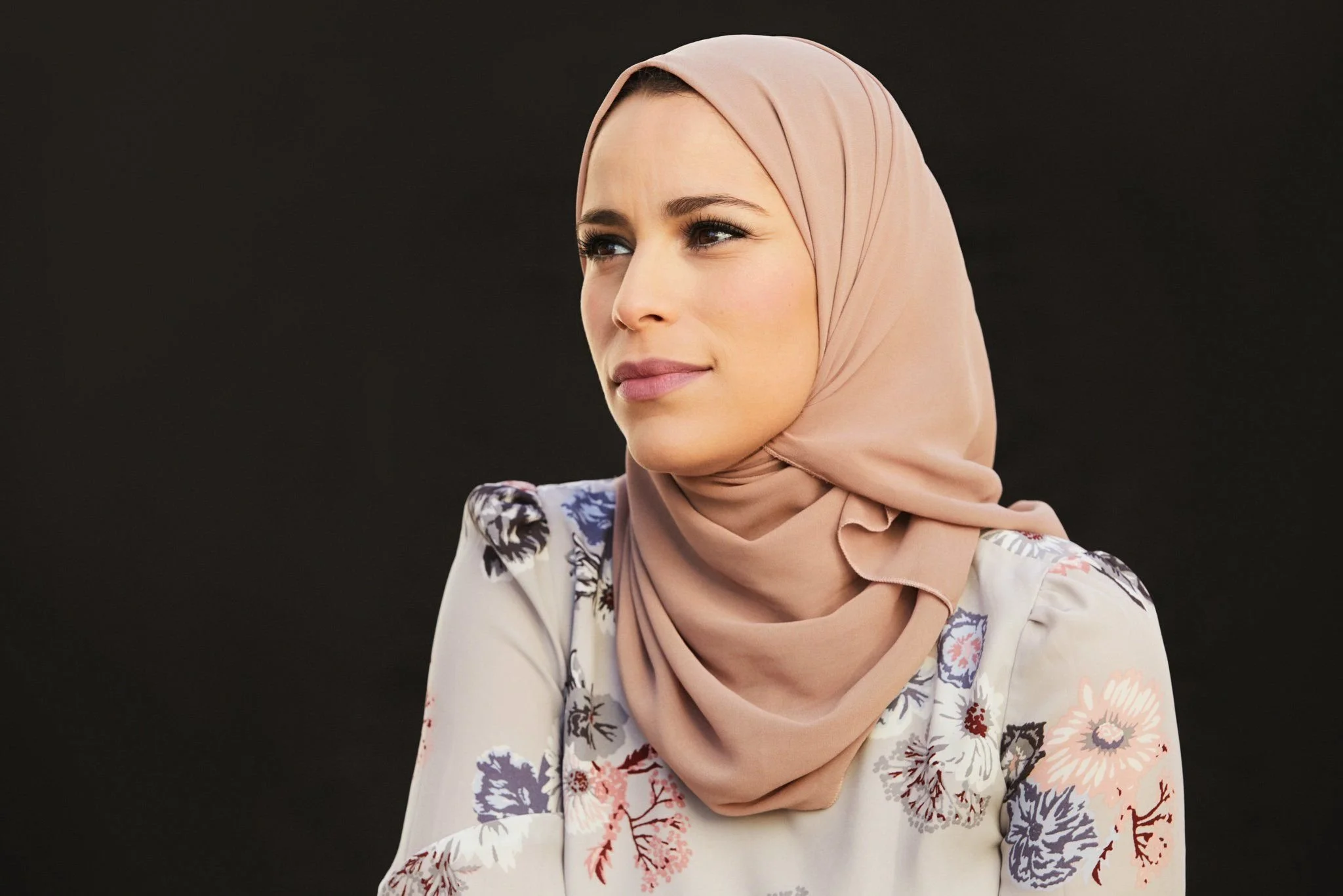 Dr. Alaa Murabit wearing a beige hijab and a floral blouse, looking to her right with a thoughtful expression, against a dark background.