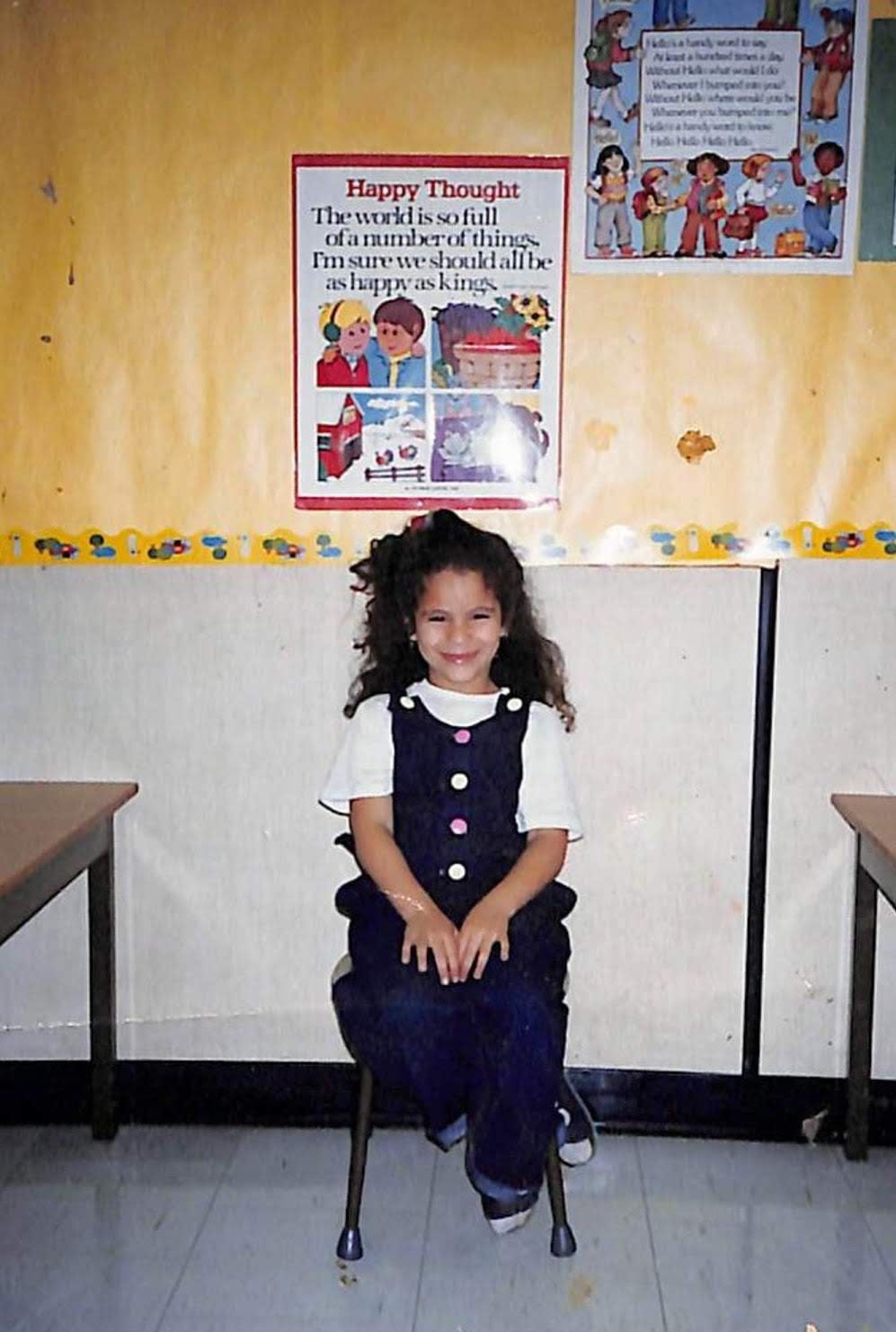 A Dr. Alaa Murabit with curly hair sitting on a chair in a classroom, smiling at the camera. Behind her is a yellow bulletin board with educational posters and colorful border decorations.