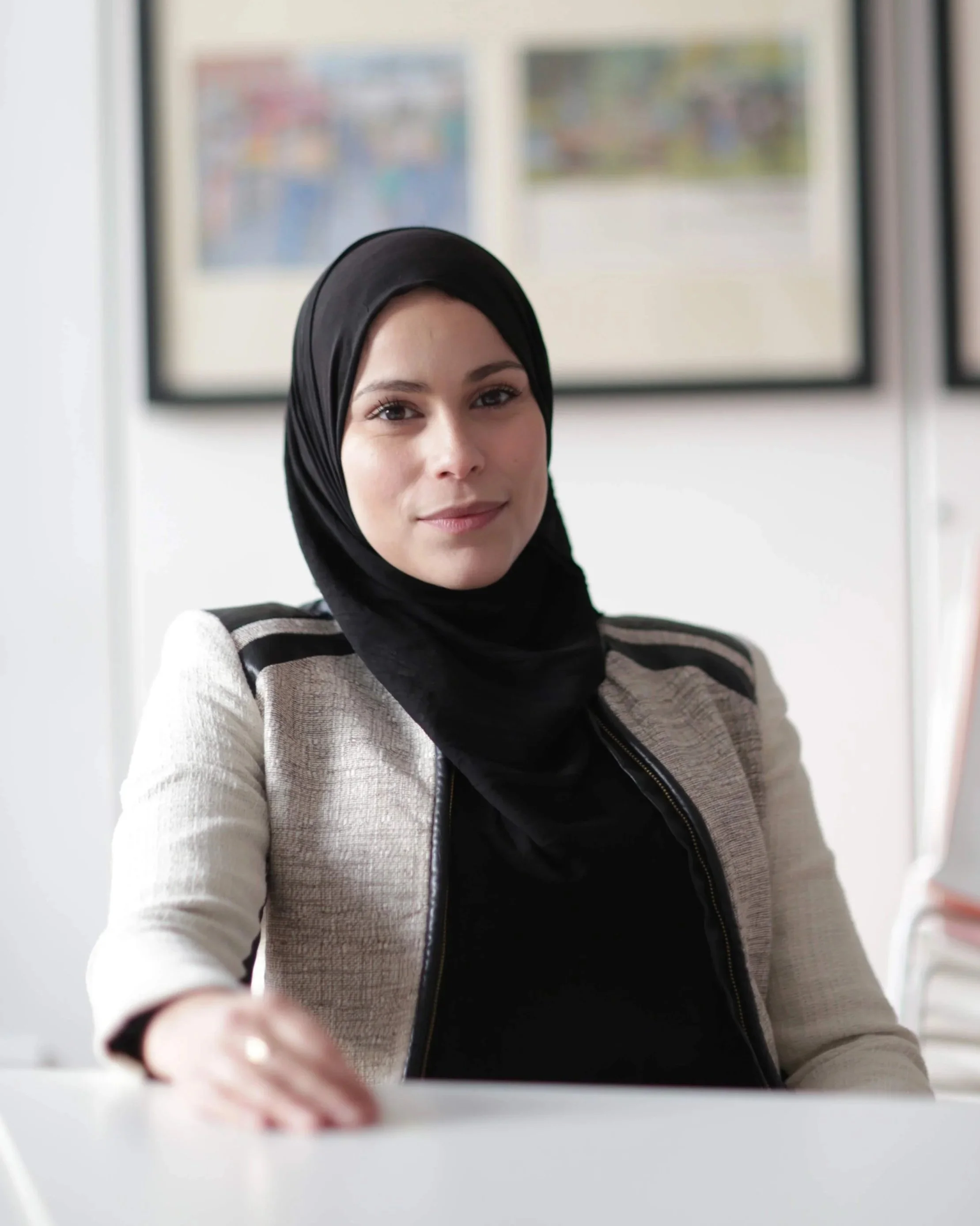 A woman wearing a black hijab and white blazer sitting at a desk in a professional setting, with framed artwork on the wall behind her.