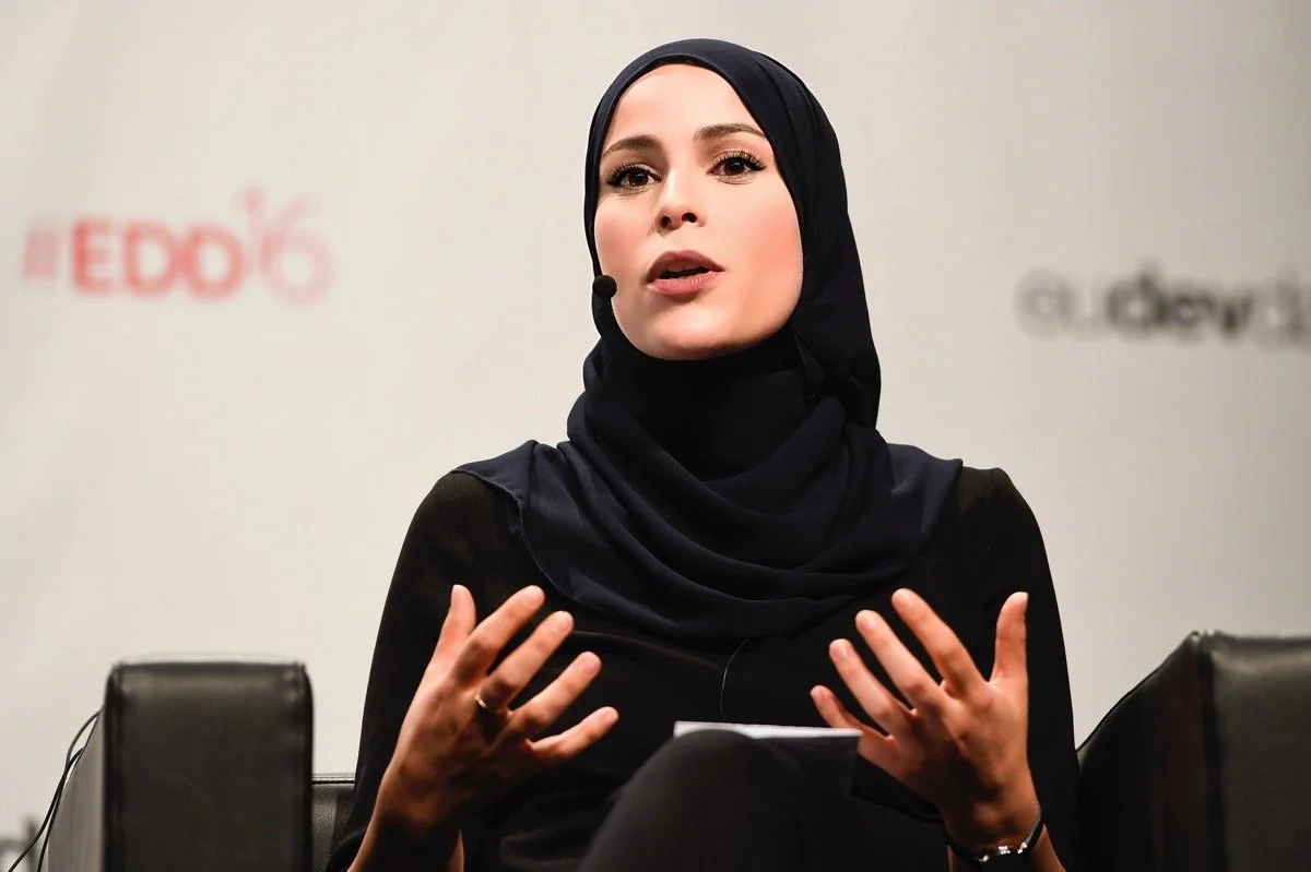 Dr. Alaa Murabit wearing a black hijab speaking at a panel discussion with AEDK6 branding in the background.