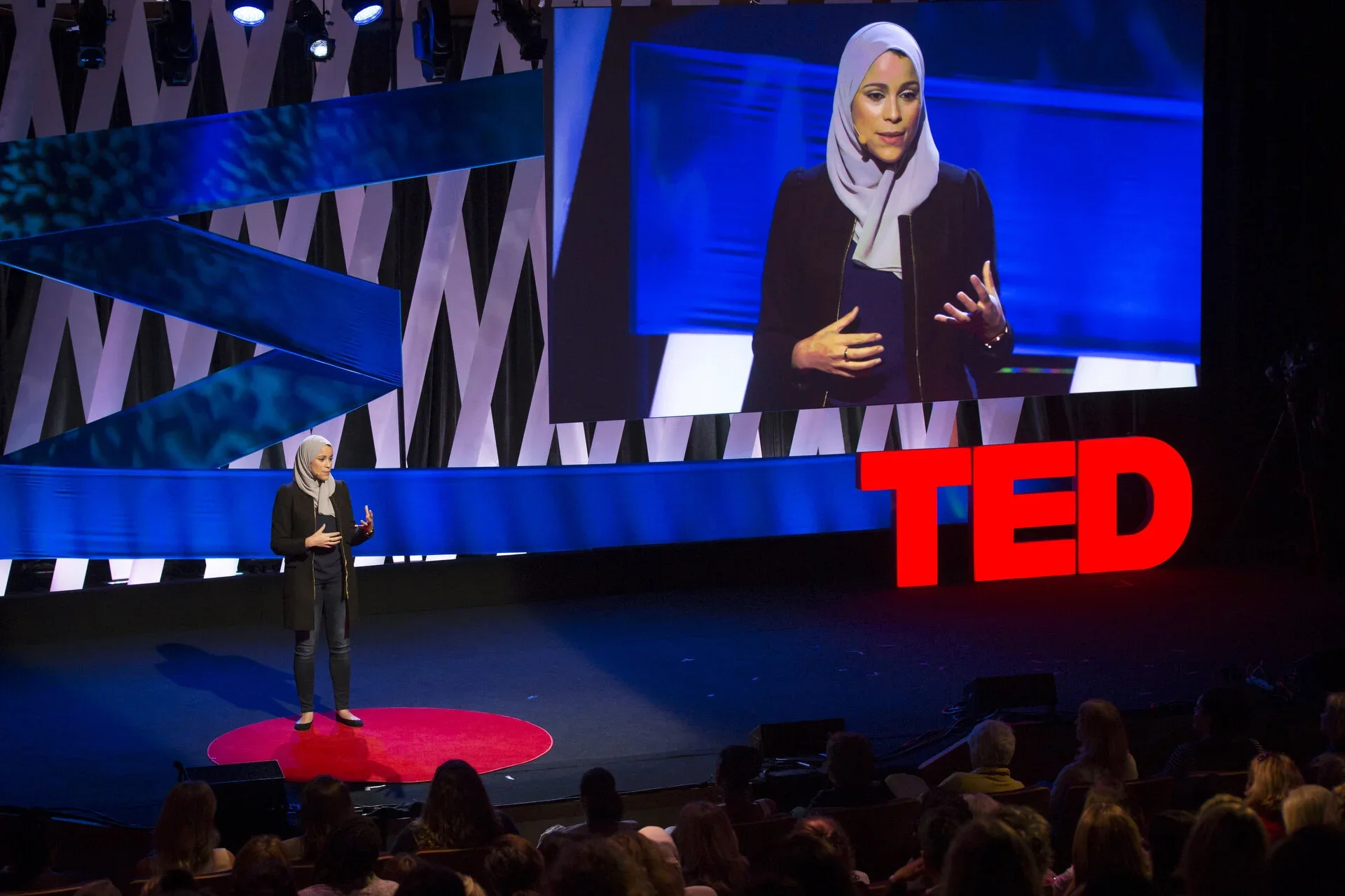 Dr. Alaa Murabit  wearing a hijab giving a TED Talk on stage, with a large screen behind her displaying her image and a red TED logo.