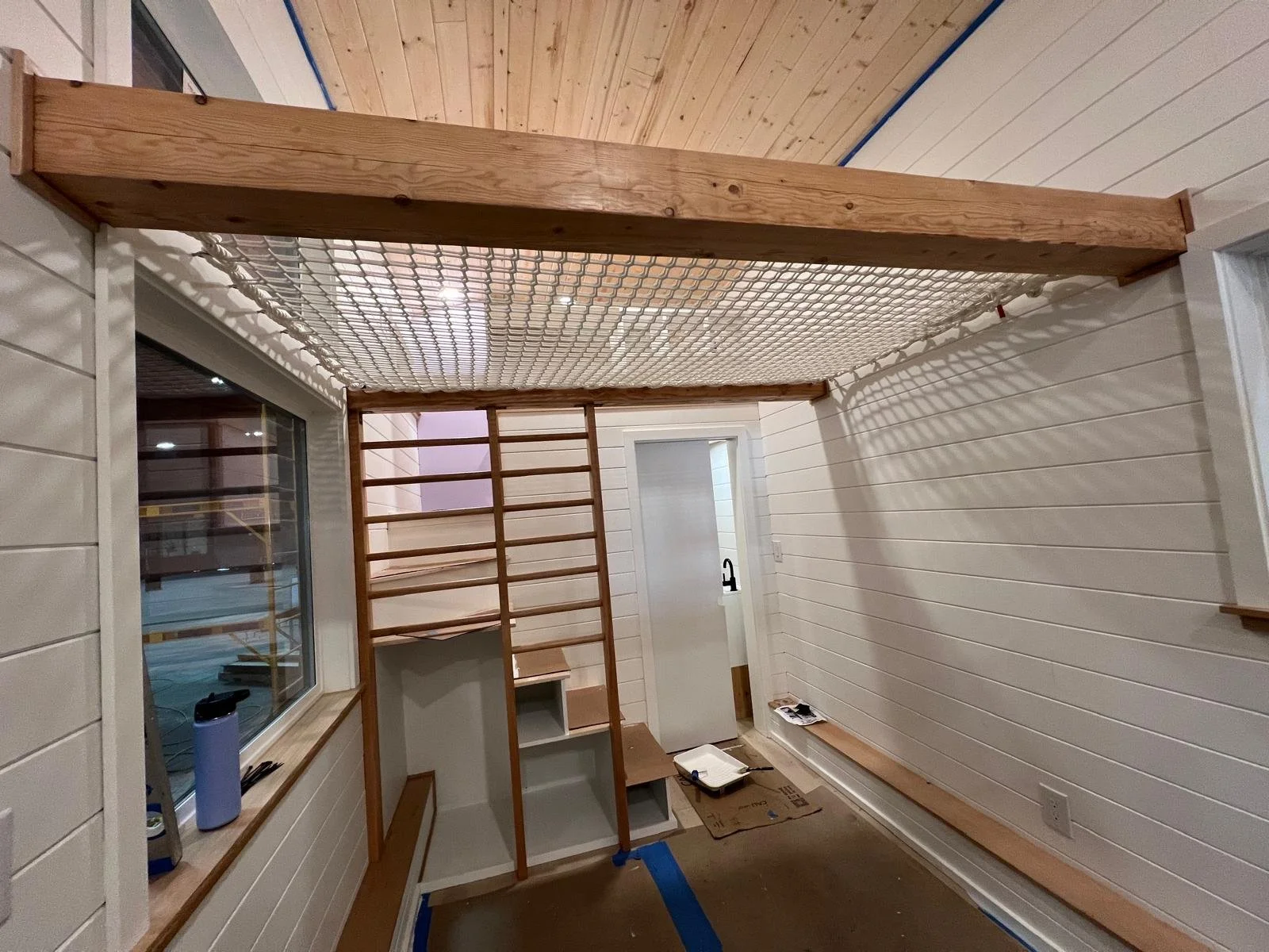 Interior of a room under construction featuring a loft with a wooden railing, a ladder, a window with a sill, a door with a black handle, and construction tools and materials on the floor.