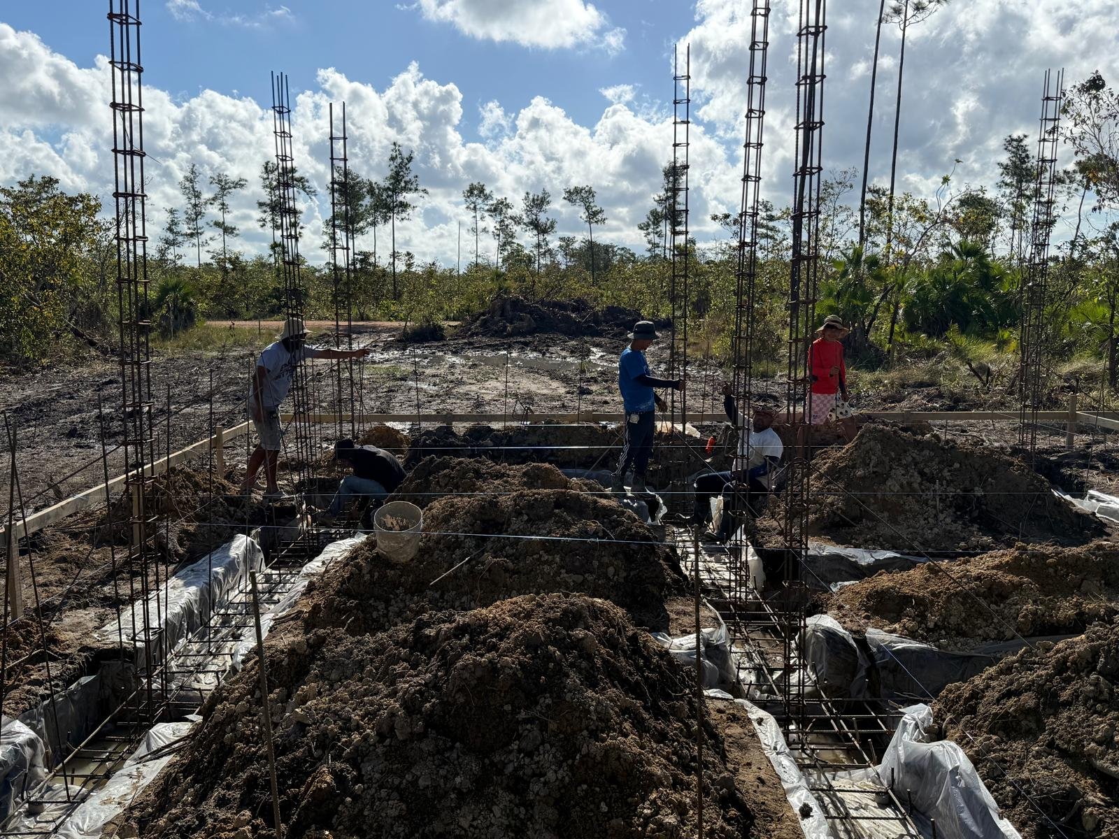 Construction workers building a foundation with rebar in a rural area surrounded by trees and cloudy sky.