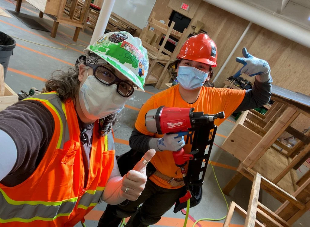 Two women construction workers in safety gear taking a selfie in a woodworking shop. One is wearing a helmet with a cartoon graphic, glasses, a mask, and a safety vest; the other is wearing an orange helmet, mask, gloves, and holding a power tool. Th