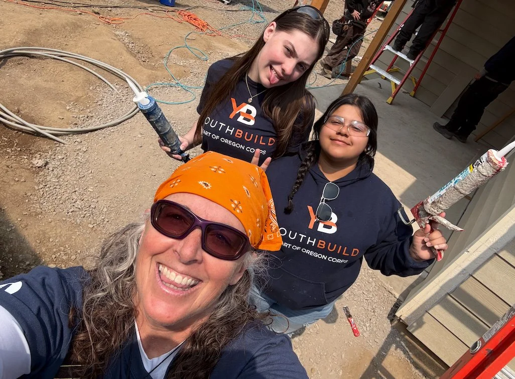 Three women at a construction site taking a selfie. They are wearing work clothes and holding caulking guns. One woman in front is wearing sunglasses and a bandana. The background includes construction tools and a partially built house.