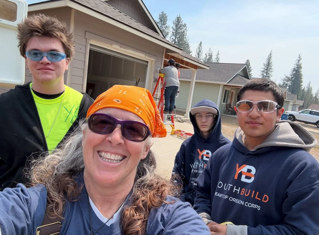 Four volunteers wearing sunglasses and hoodies, standing outdoors in front of a house under construction, with a person working on the house in the background.
