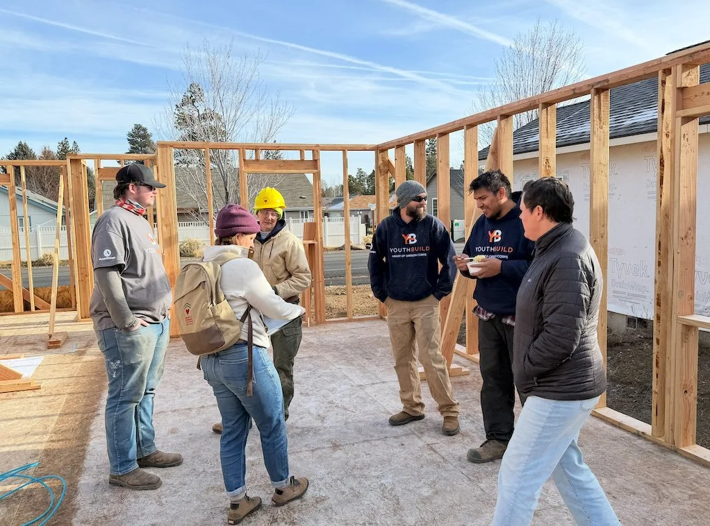 Group of six people at a construction site, some wearing safety gear, discussing next steps as a wooden house frame is being built on a clear day.
