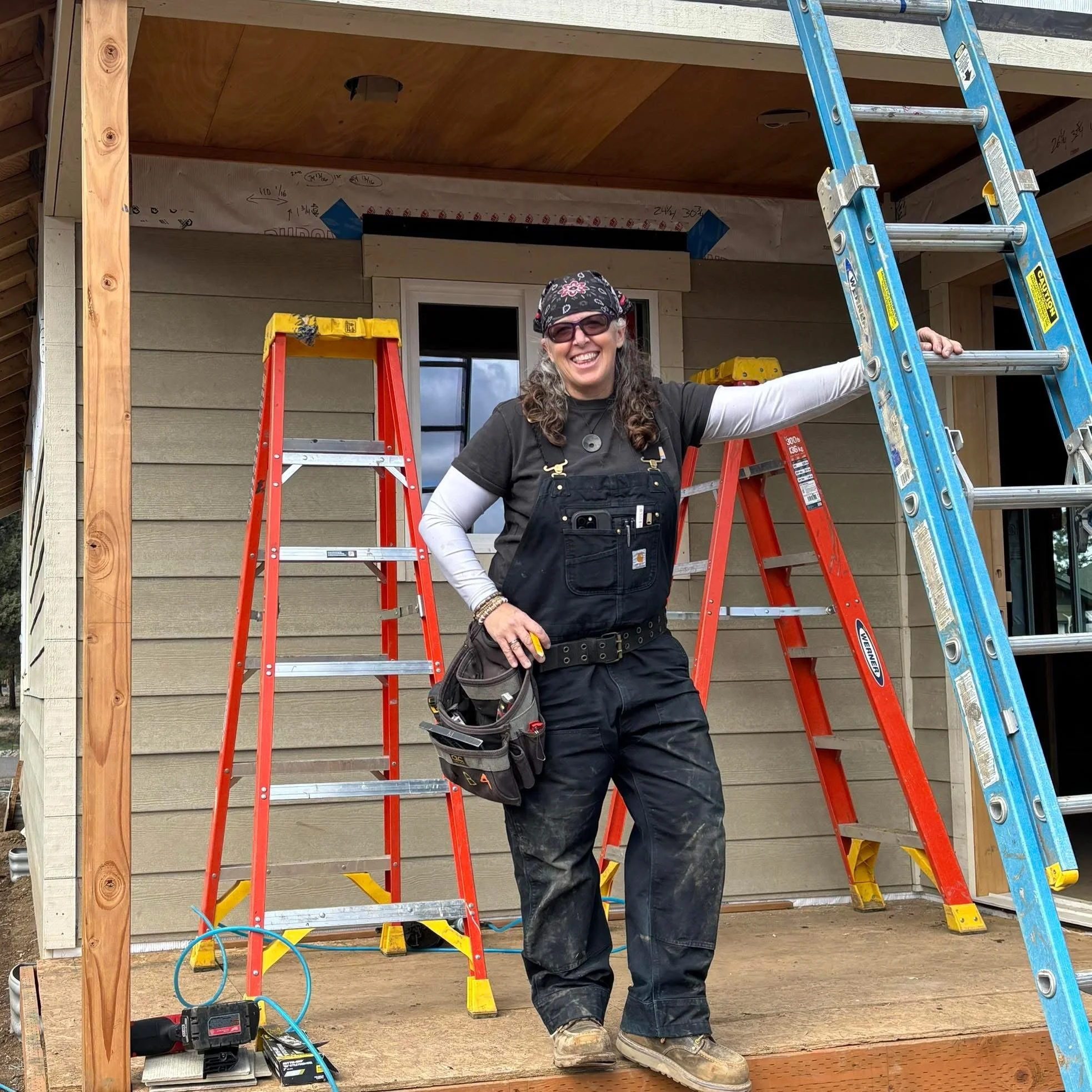 A woman on a construction site smiling, wearing safety glasses, a bandana, a black shirt, and overalls. She is standing next to a ladder and a partially built house with wooden framing and siding.