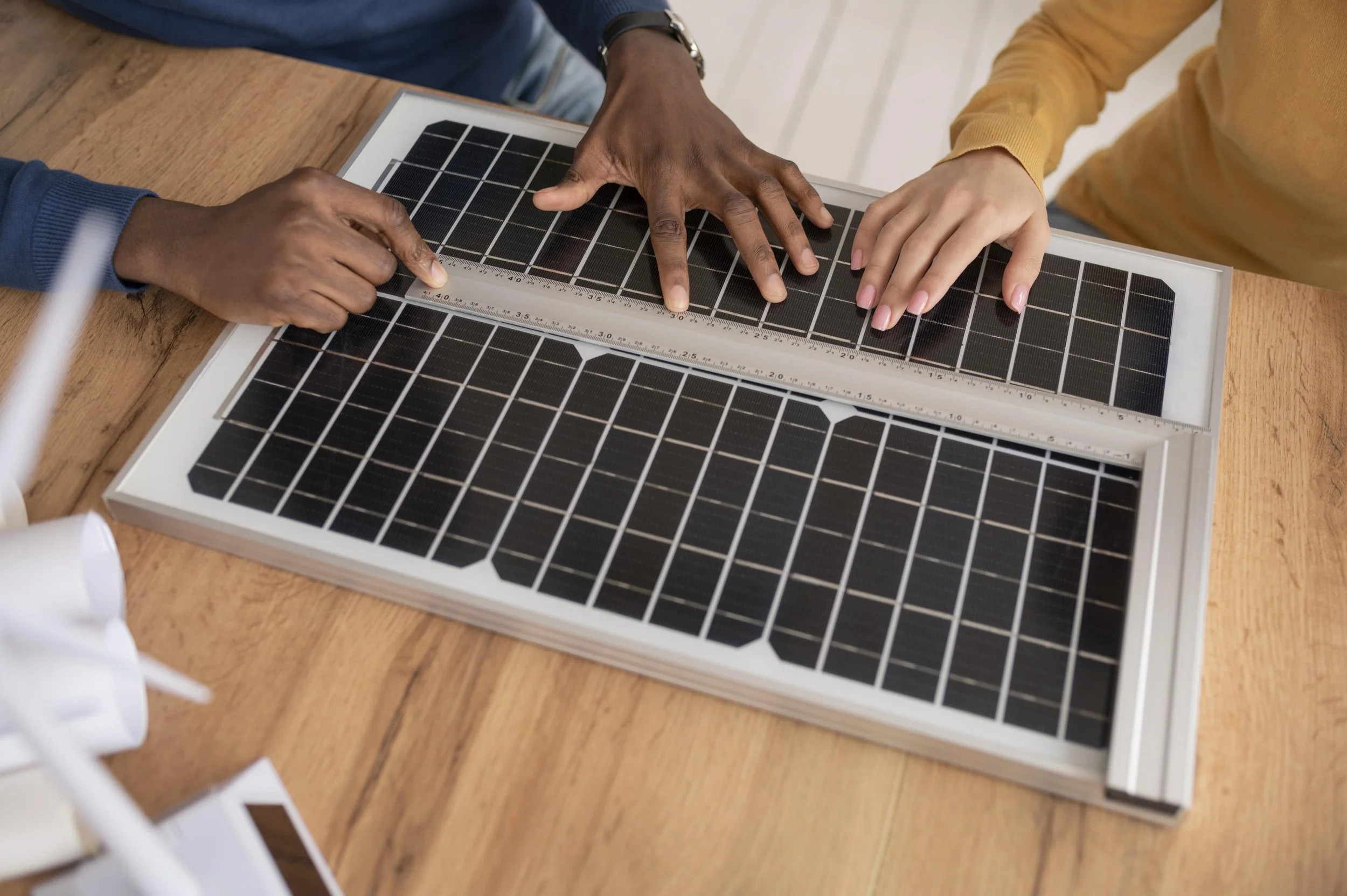 Hands of two people using a ruler to measure a solar panel on a table.