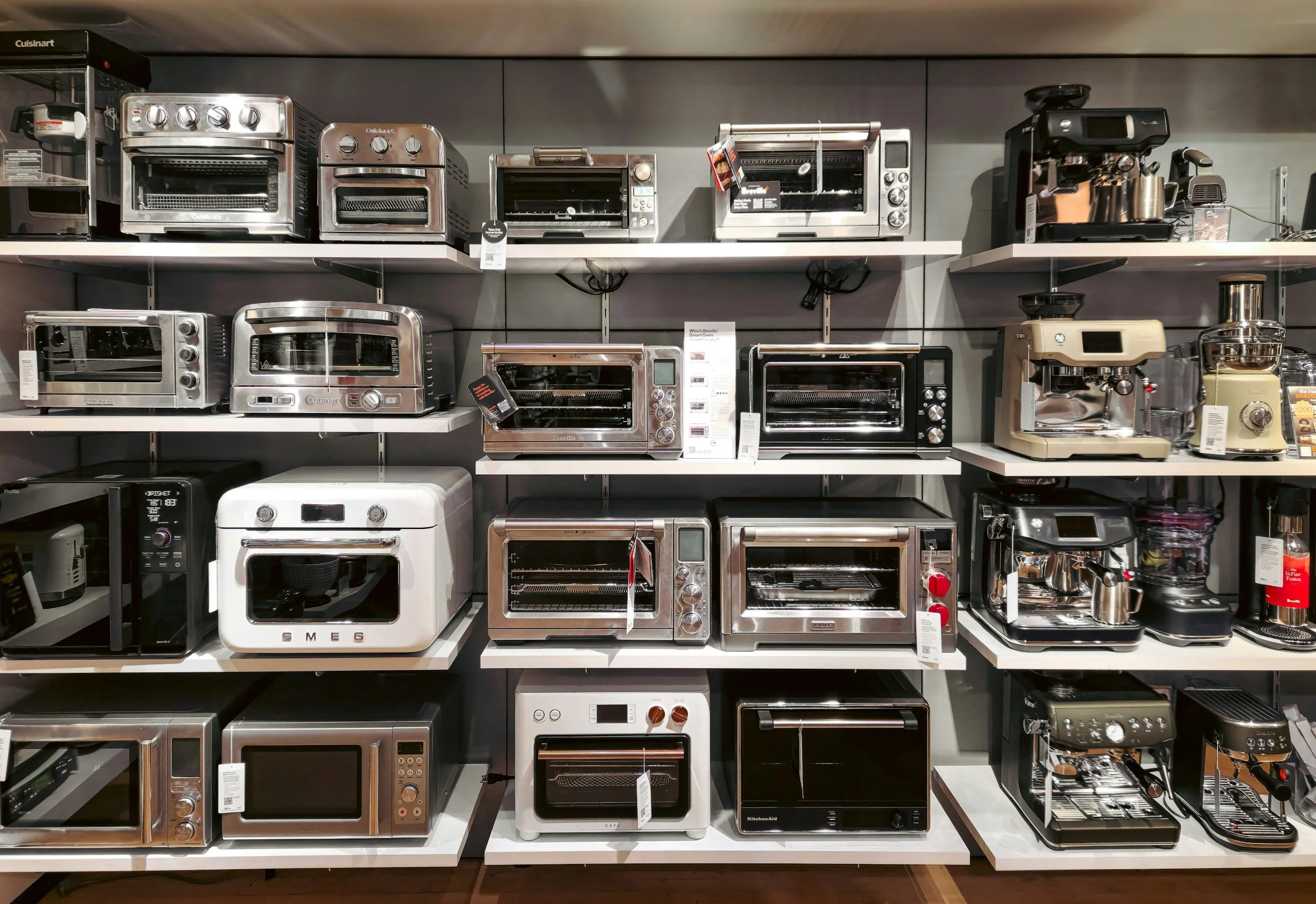 A store display shelf filled with various toasters, bread ovens, and kitchen appliances, primarily in stainless steel and black finishes.