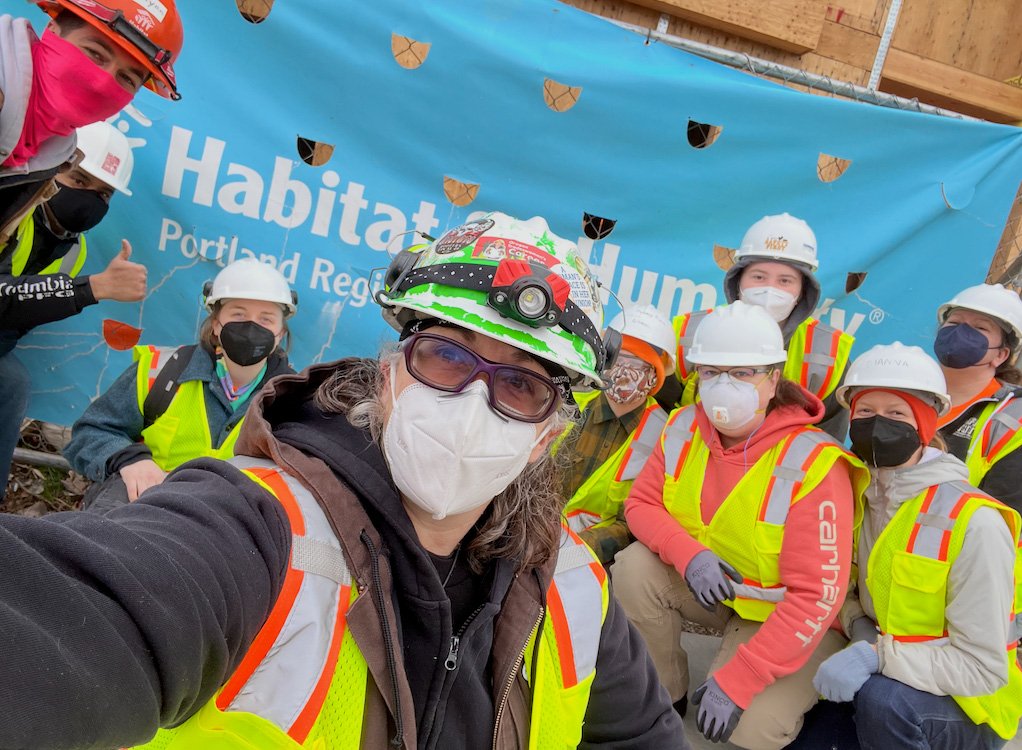 Group of people in construction gear, hard hats, and masks posing for a selfie at Habitat for Humanity project site.