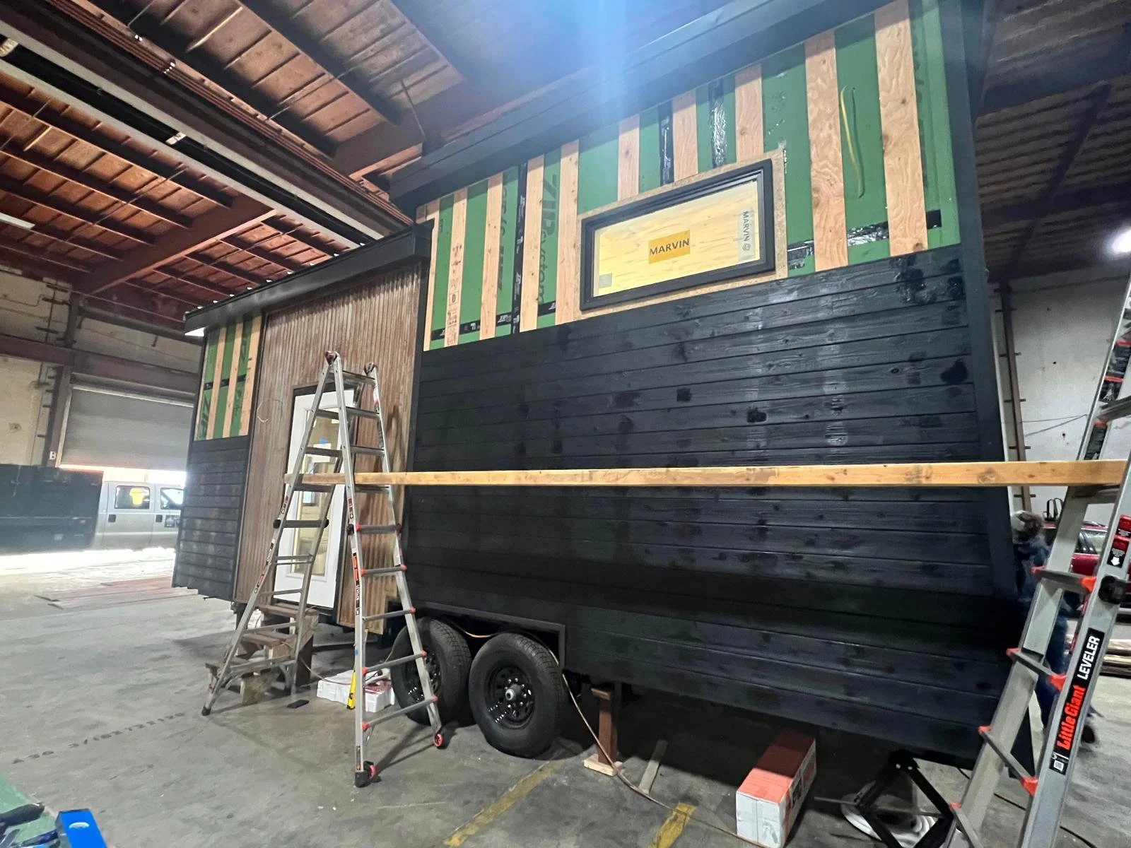 Inside a warehouse, a trailer is being converted into a tiny house with black and green siding, with ladders, tools, and construction materials around.