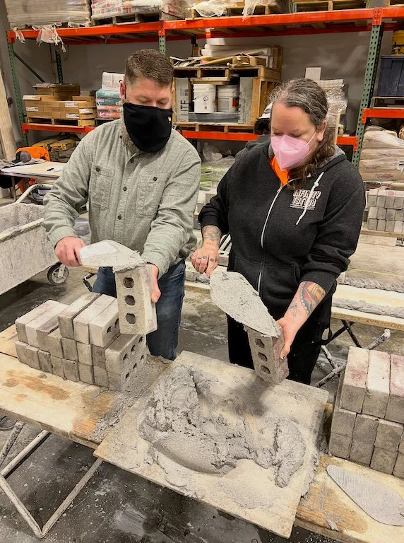 Two people wearing masks are stacking concrete blocks and working with a mold, possibly for cement or concrete artwork, in a warehouse or workshop setting.