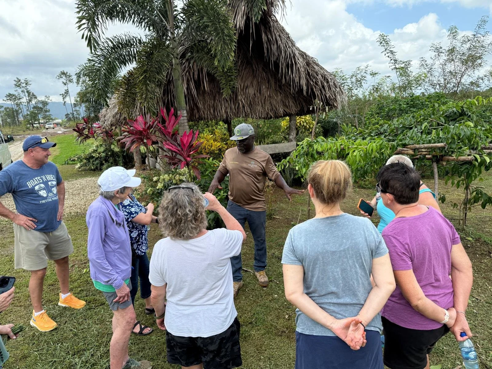 A group of people gathered outdoors around a guide giving a presentation near a thatched-roof hut, with tropical plants and trees in the background.