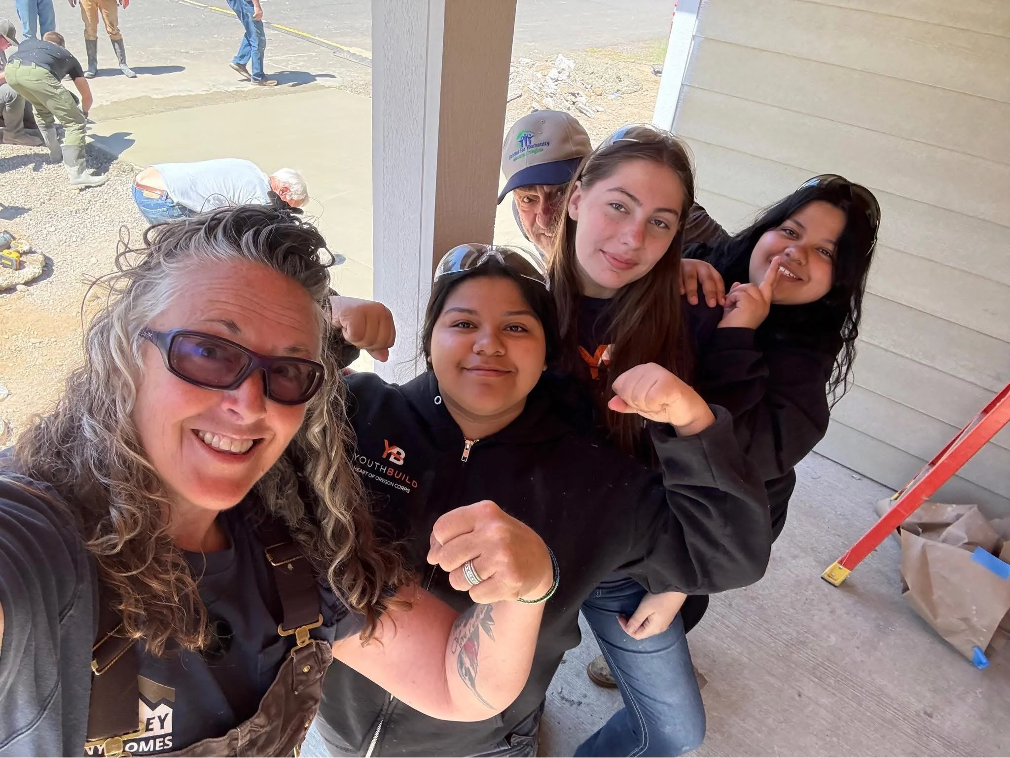 Smiling group of diverse people taking a selfie together at a construction site. Background shows workers and construction materials.