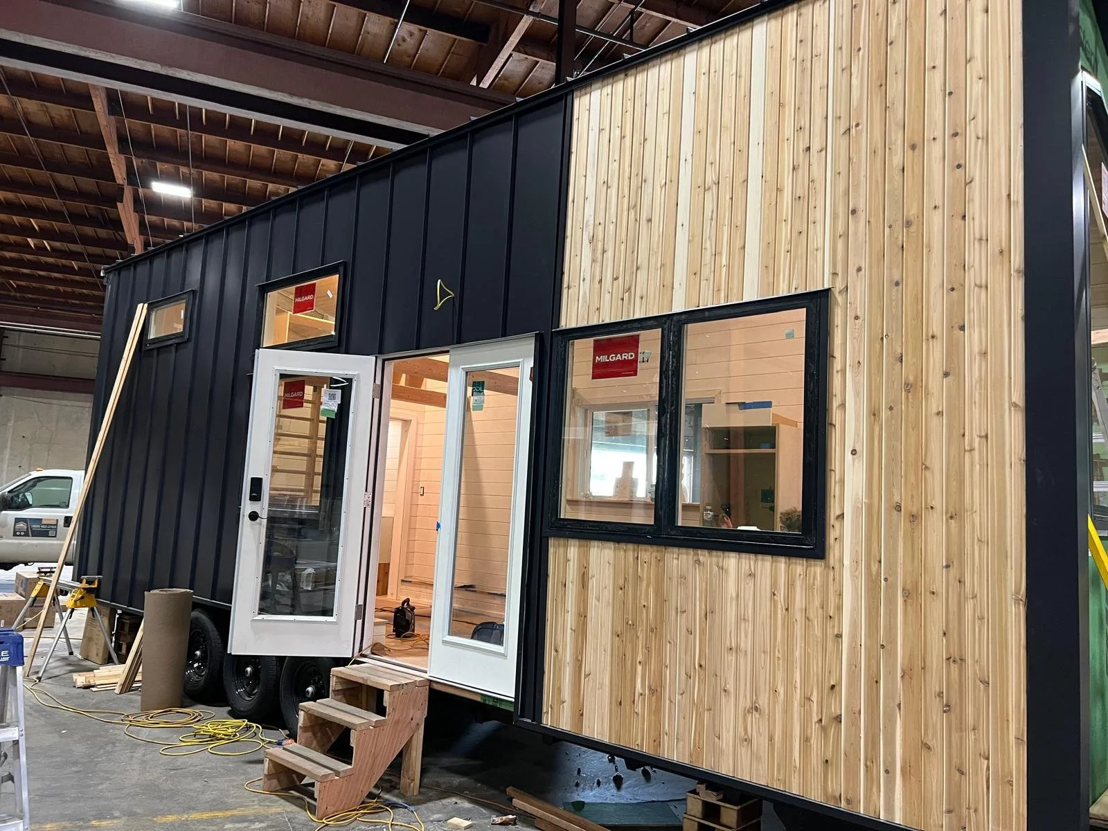 A tiny house under construction inside a large indoor facility, with a black and wood exterior, partially open door, and windows showing the interior. Construction tools and materials are nearby.