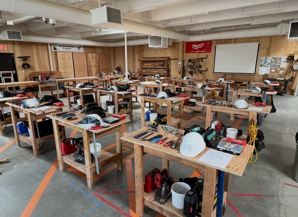 Workshop classroom with wooden workbenches arranged in rows, each with safety helmets, tools, and safety gear, set up for a woodworking or construction training session.