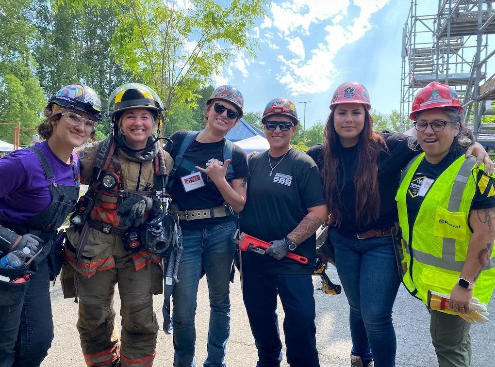 A group of six women dressed in safety gear, including helmets and vests, standing outdoors with trees and blue sky in the background.