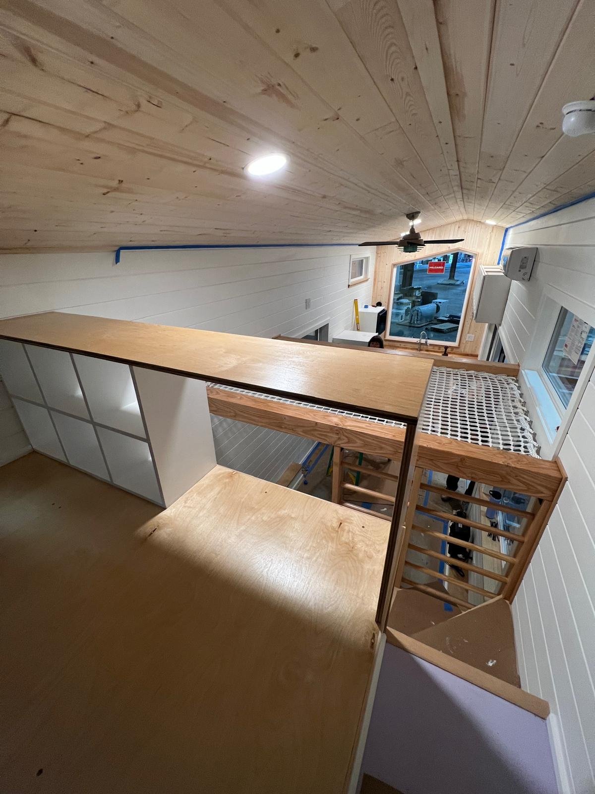 Interior of a house under construction showing a loft area with a wooden counter, stairs leading down, a ceiling fan, and large windows.