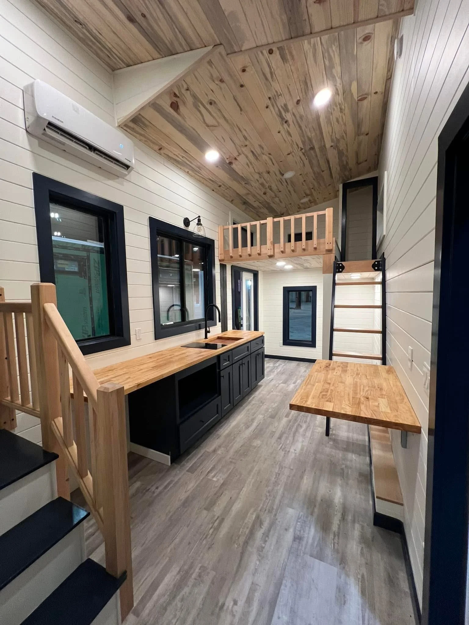 Interior of a modern, rustic-style kitchen with wooden ceiling and white shiplap walls. Black-framed windows, a black kitchen cabinet with a wooden countertop, a black faucet, and a small wooden table. A ladder leads up to a small loft area with a wo