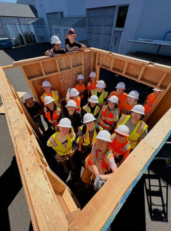 Group of children and adults wearing safety vests and hard hats inside a wooden construction frame, possibly on a construction site.