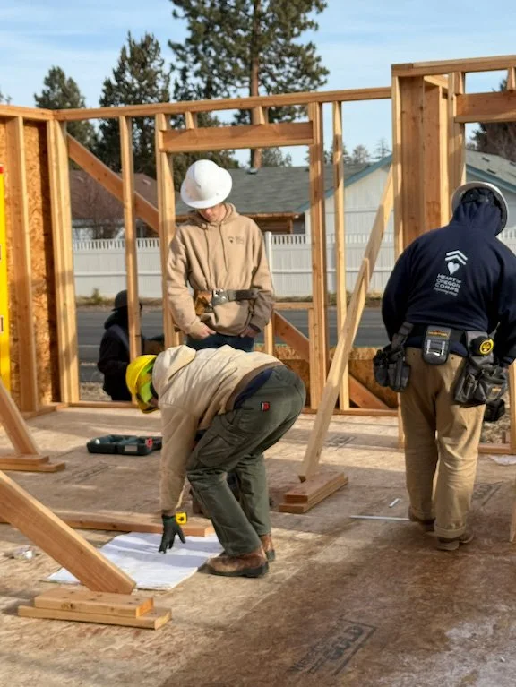 Construction workers assembling a wooden frame for a building on a construction site outdoors with trees and houses in the background.