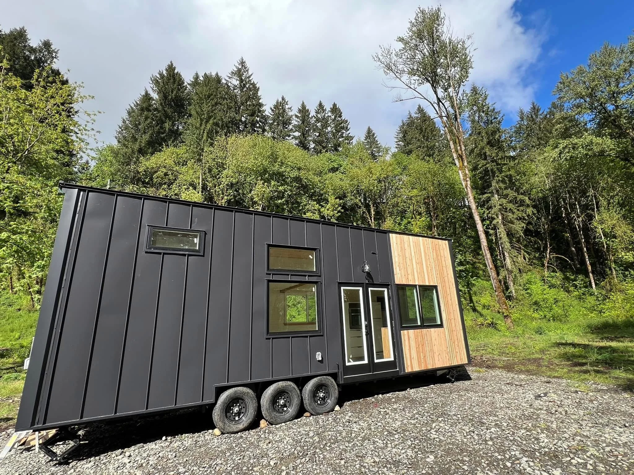 A modern tiny house with black metal siding and wooden accent wall, resting on a trailer, surrounded by trees and a partly cloudy sky.
