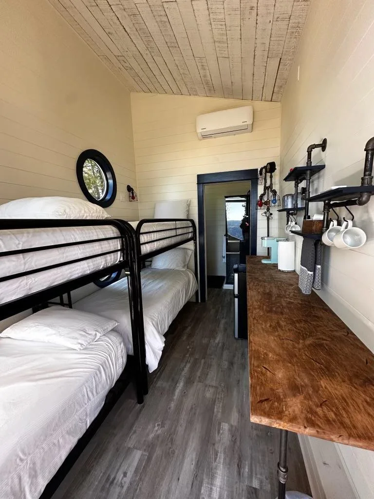 A small bedroom with black metal bunk beds, white bedding, a round window, a wooden dresser with hanging mugs, a small white appliance, and a doorway leading outside, with beige walls and a wooden ceiling.