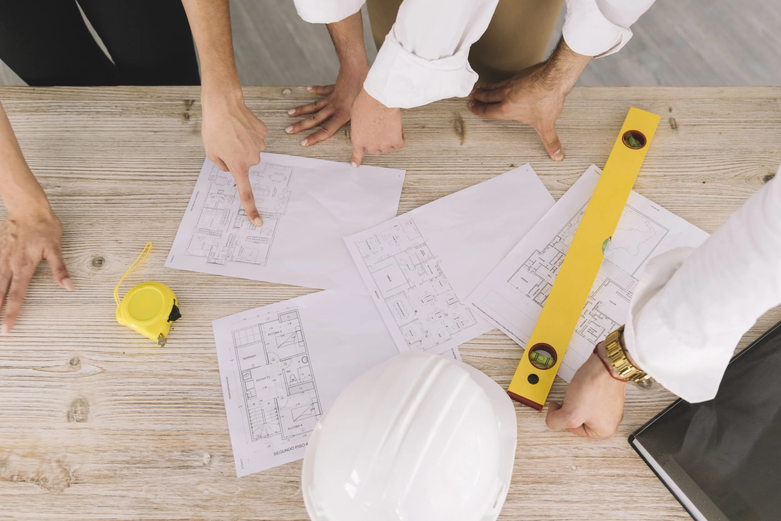 People reviewing blueprints and architectural plans on a wooden table, with a yellow spirit level, a yellow tape measure, and a tablet visible.