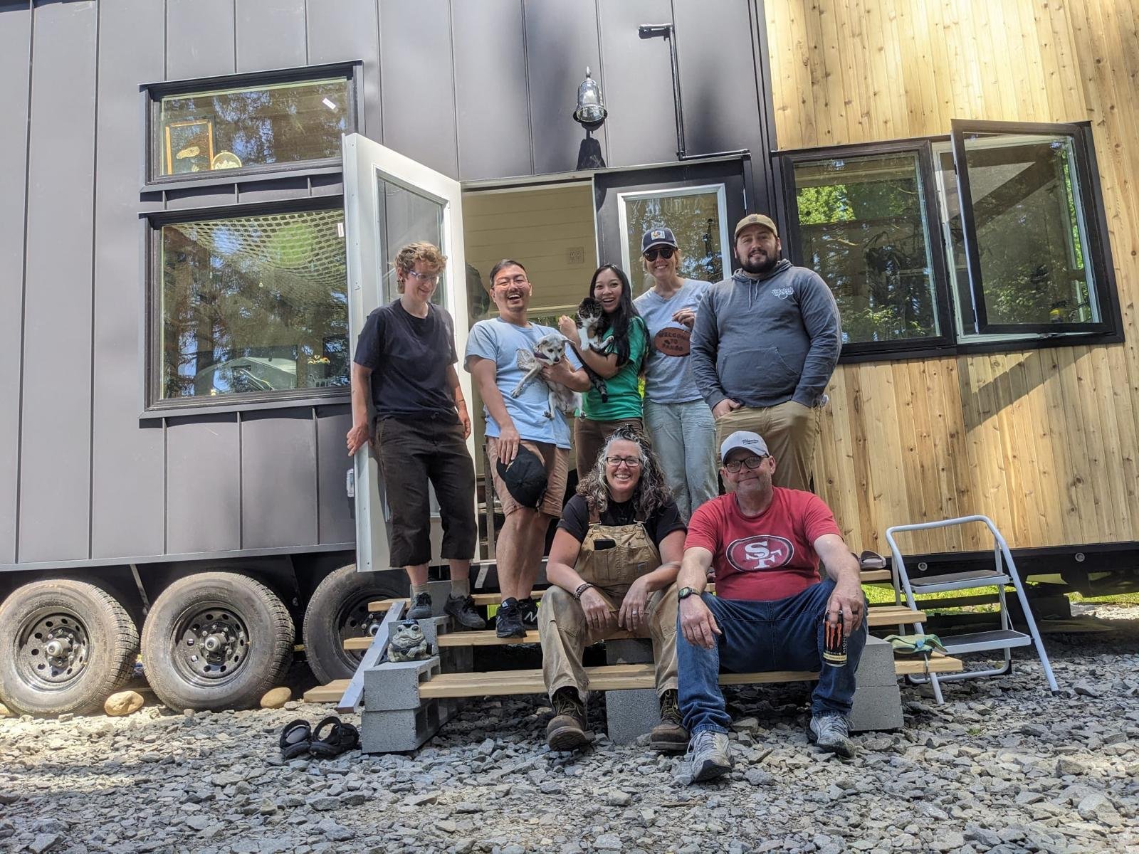 Group of people standing and sitting on front steps in front of tiny house on wheels, surrounded by trees and rocks, smiling and holding two dogs, with open windows and a wooden exterior.