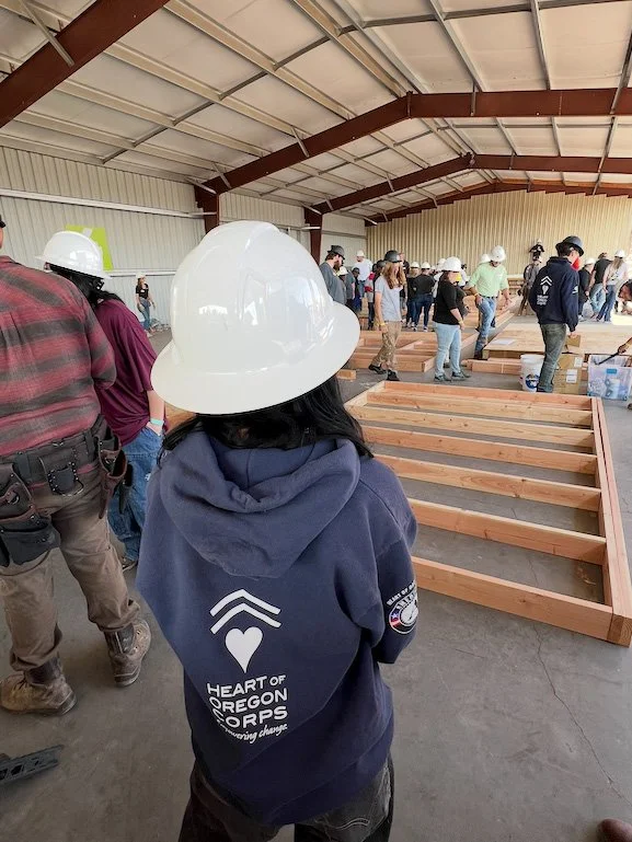 Group of people in a warehouse or workshop building with high ceiling, wearing safety helmets, gathered around partially assembled wooden bed frames.
