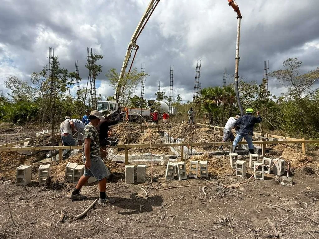 Construction workers laying the foundation for a building, pouring concrete, and setting cinder blocks on a muddy site with steel reinforcement bars and a crane overhead under cloudy skies.