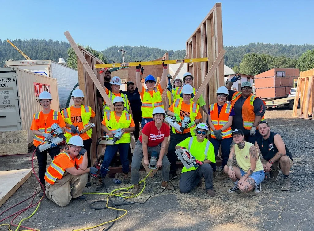 Group of construction workers and volunteers wearing safety vests and hard hats, working together on a building frame at a construction site.