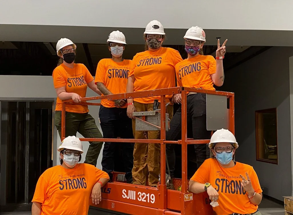 Group of six women construction workers wearing orange shirts with 'STRONG' printed on them, white hard hats, and face masks, standing on a construction lift inside a building, with some women giving peace signs and one making a rock gesture.