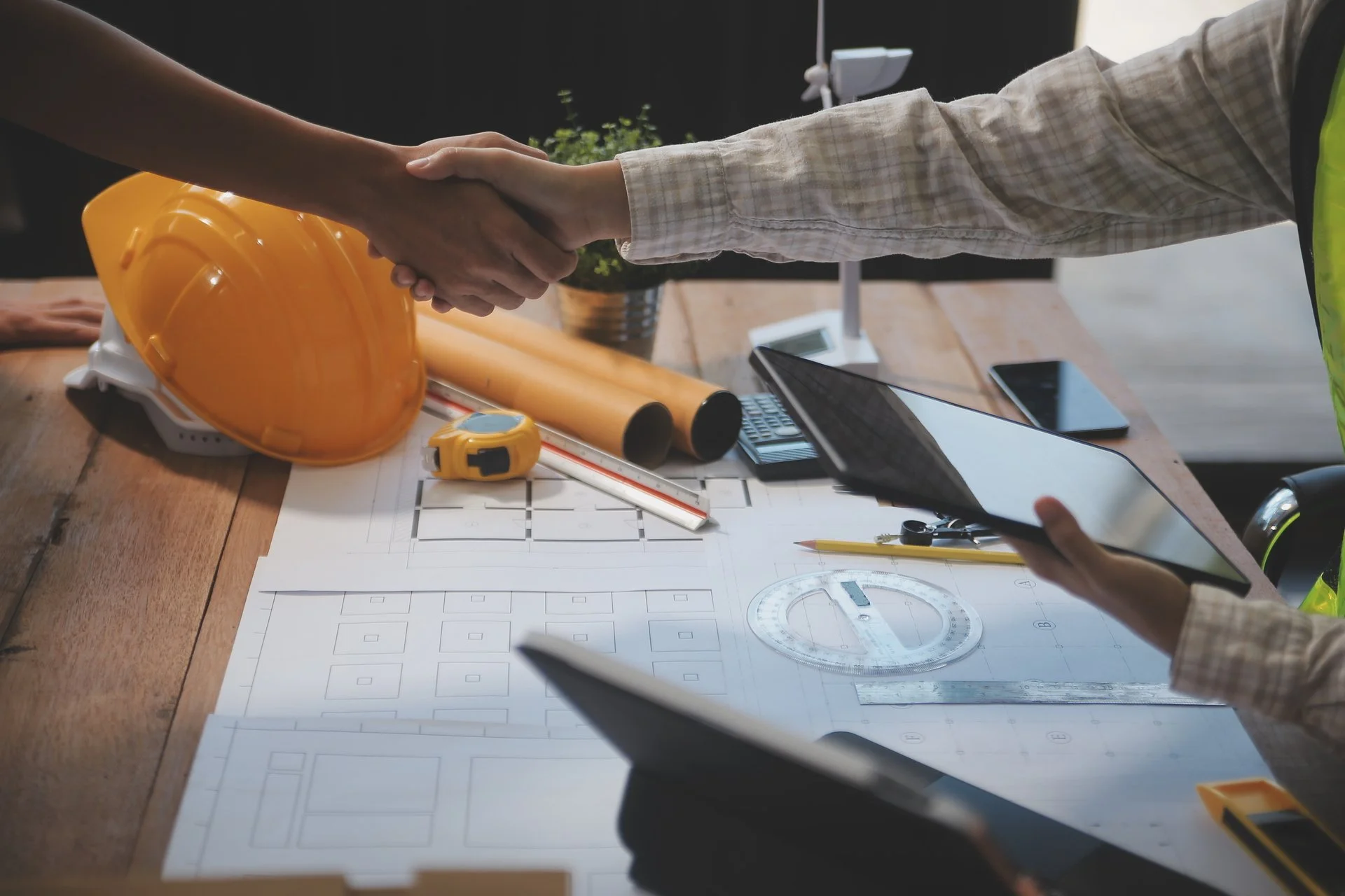 Handshake between two people in a construction office, with a yellow safety helmet, measuring tape, and architectural plans on the table.