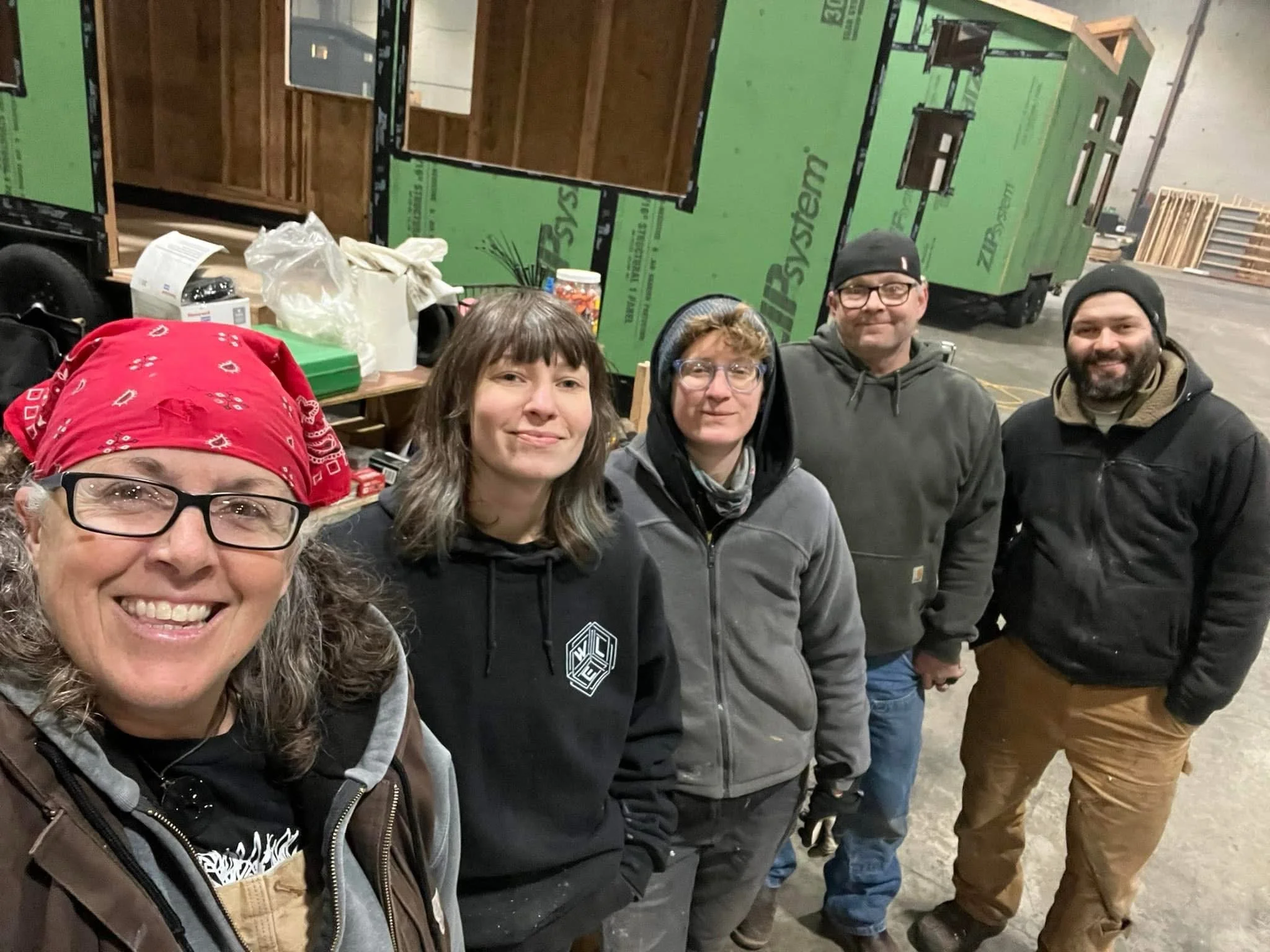 Group of five people standing in a row inside a warehouse or workshop, with green insulation panels and wooden pallets in the background.