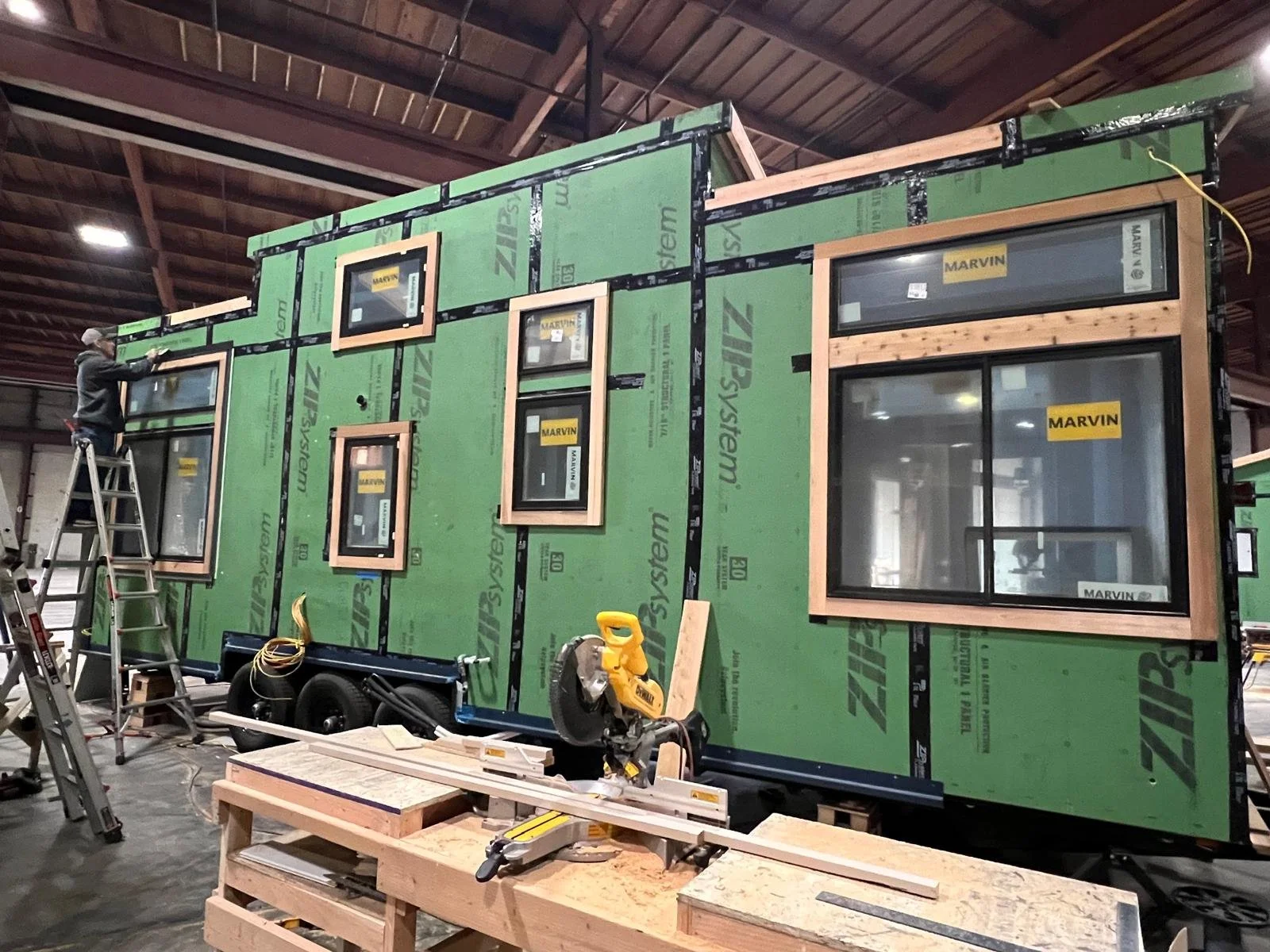 Construction of a tiny house with green sheathing, black trim, and multiple windows, inside a large warehouse