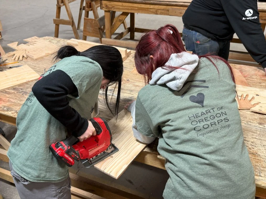 Two people working on a woodworking project in a workshop. One is using a red power saw on a piece of wood, while the other is closely observing. Both are wearing gray shirts with 'Heart of Oregon Corps' logo.