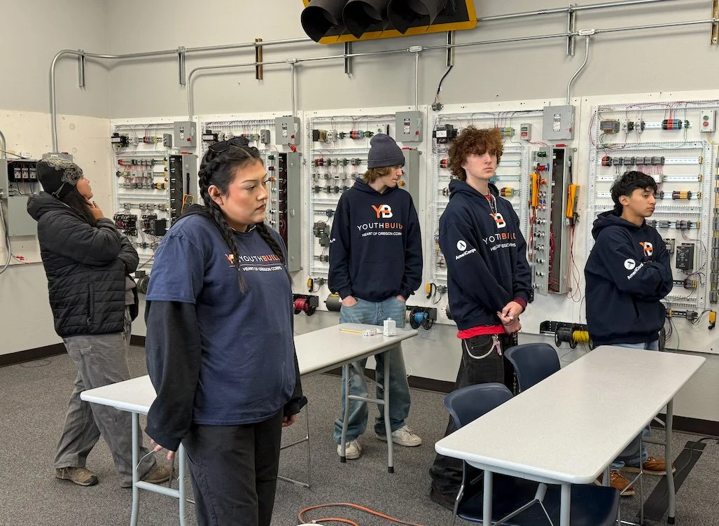 Four students and one adult in a classroom with electrical wiring and control panels, participating in a youth build program.
