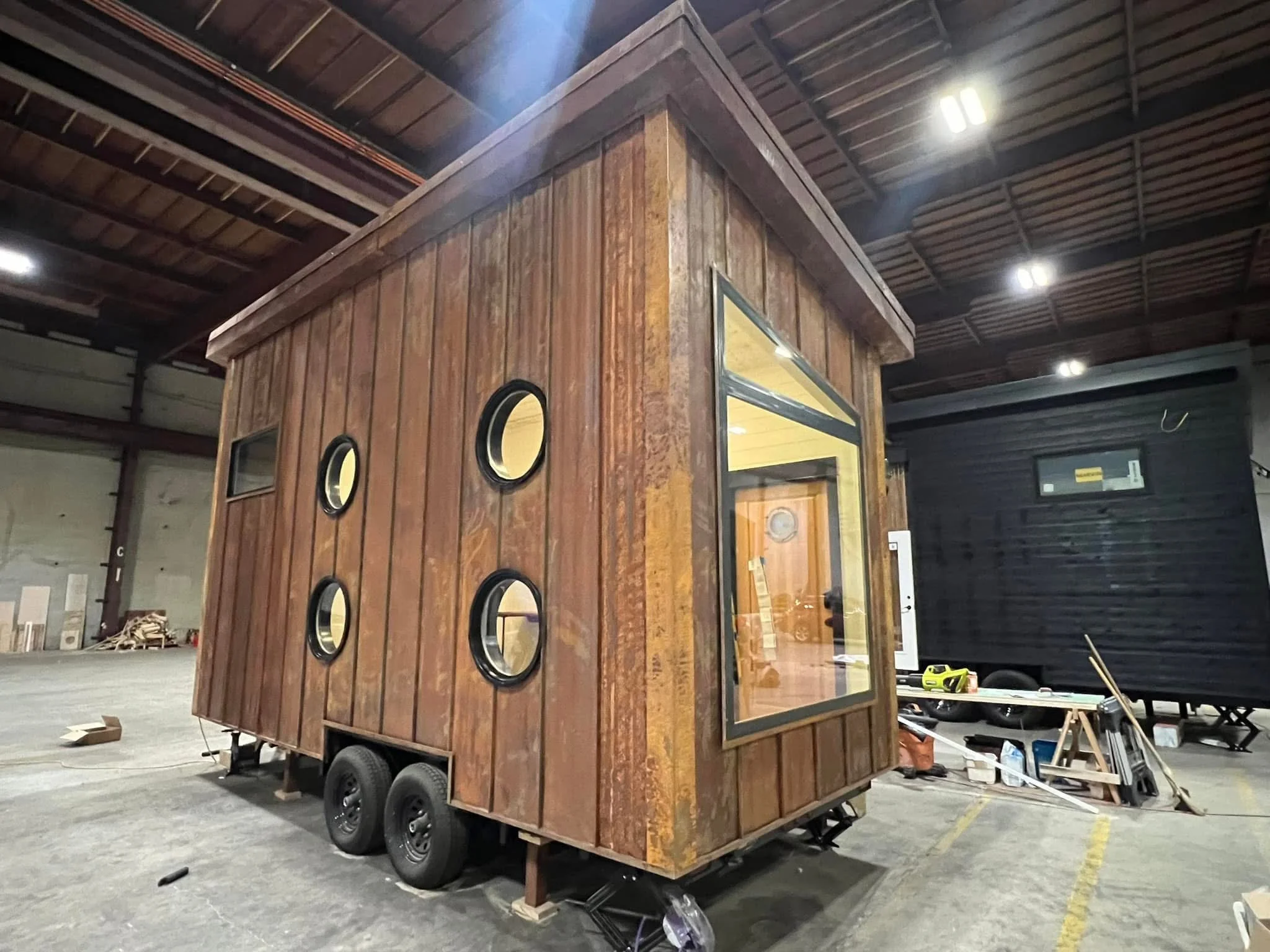 A wooden tiny house on wheels inside a warehouse with a high ceiling, fluorescent lights, and some tools and materials scattered around.