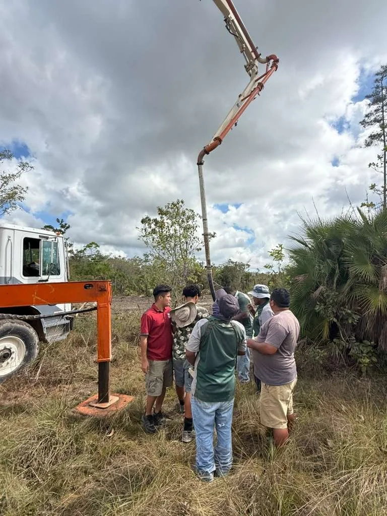 Group of people gathered around a construction truck with a long boom arm and a bucket lift, in a grassy outdoor area with trees and cloudy sky.