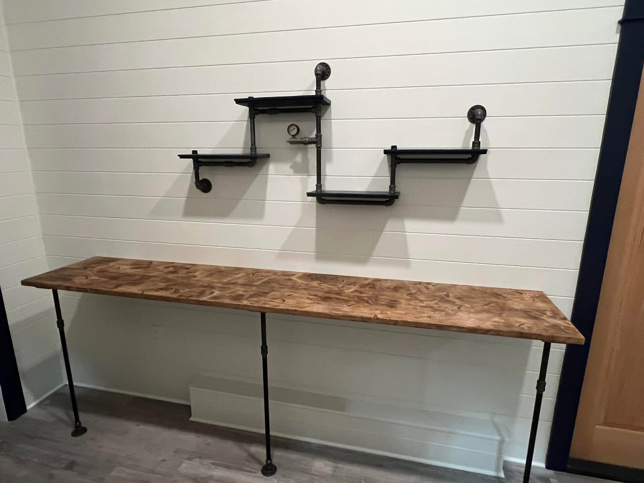 Wall-mounted black pipe shelving with three levels, a barometer, and pipe fixtures, against white shiplap walls, above a wooden table with black metal legs.