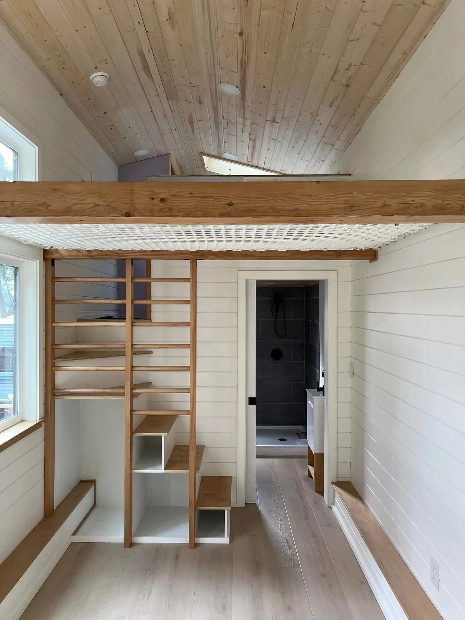 Room with light wood floors and wall paneling, a wooden loft structure under construction, and a doorway leading to a dark tiled shower area.