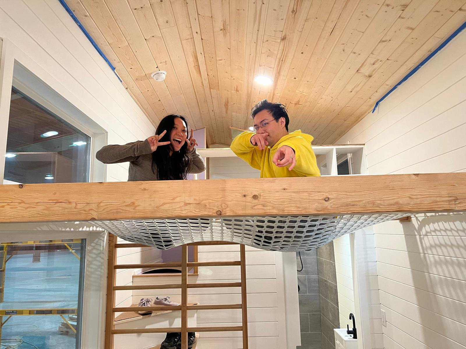 Two people standing on a wooden platform inside a home, smiling and making playful gestures toward the camera.