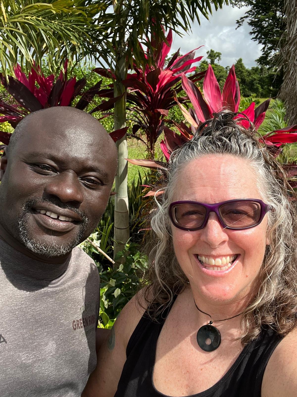 Close-up of a smiling Black man and a smiling white woman with curly hair and purple glasses taking a selfie outdoors with tropical plants and pink flowers in the background.