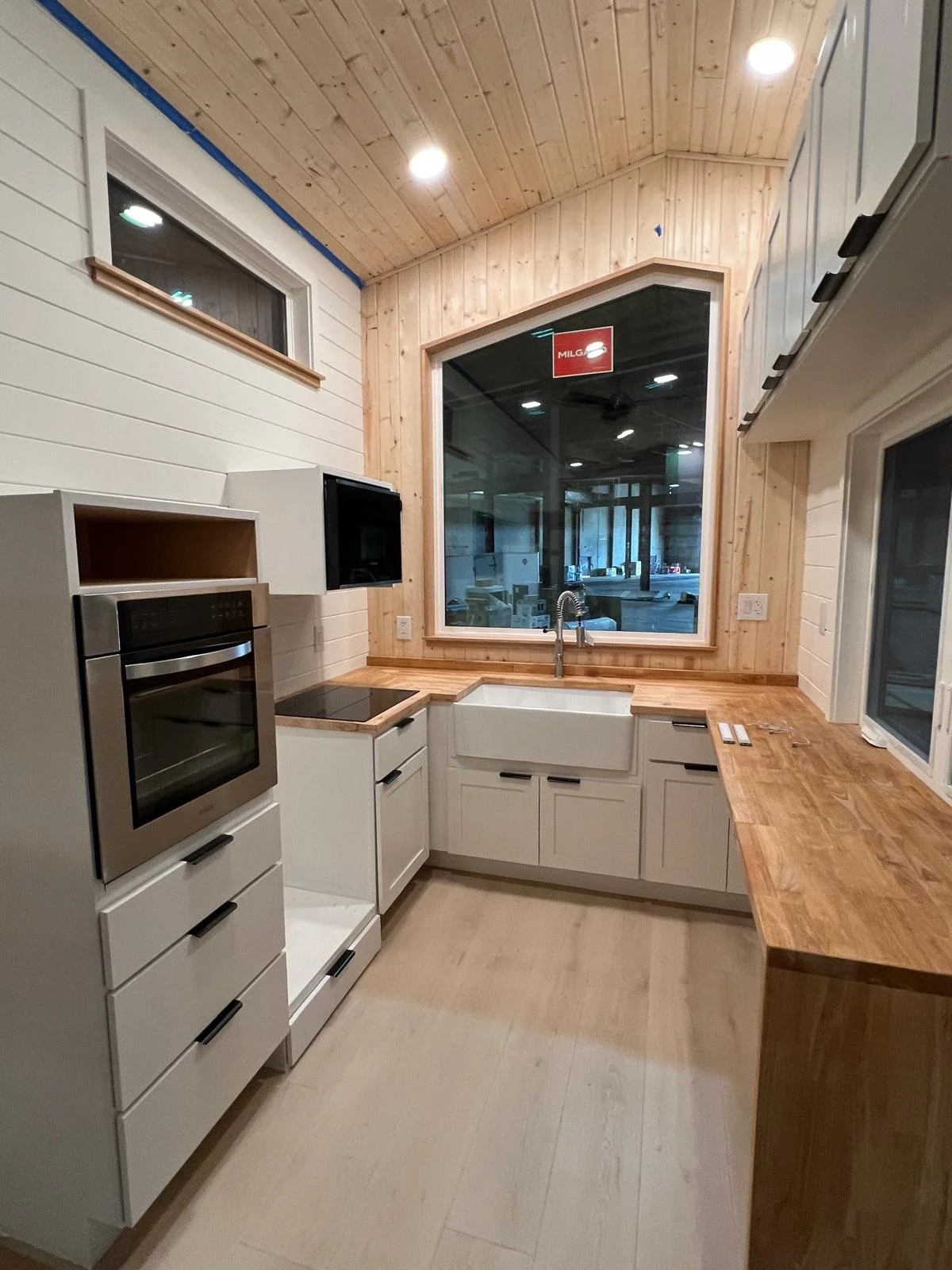 Interior view of a modern kitchen with white cabinets, wooden countertops, a farmhouse sink, and large windows showing an outdoor scene. The ceiling is wood-paneled, and there's a small window on the left wall.