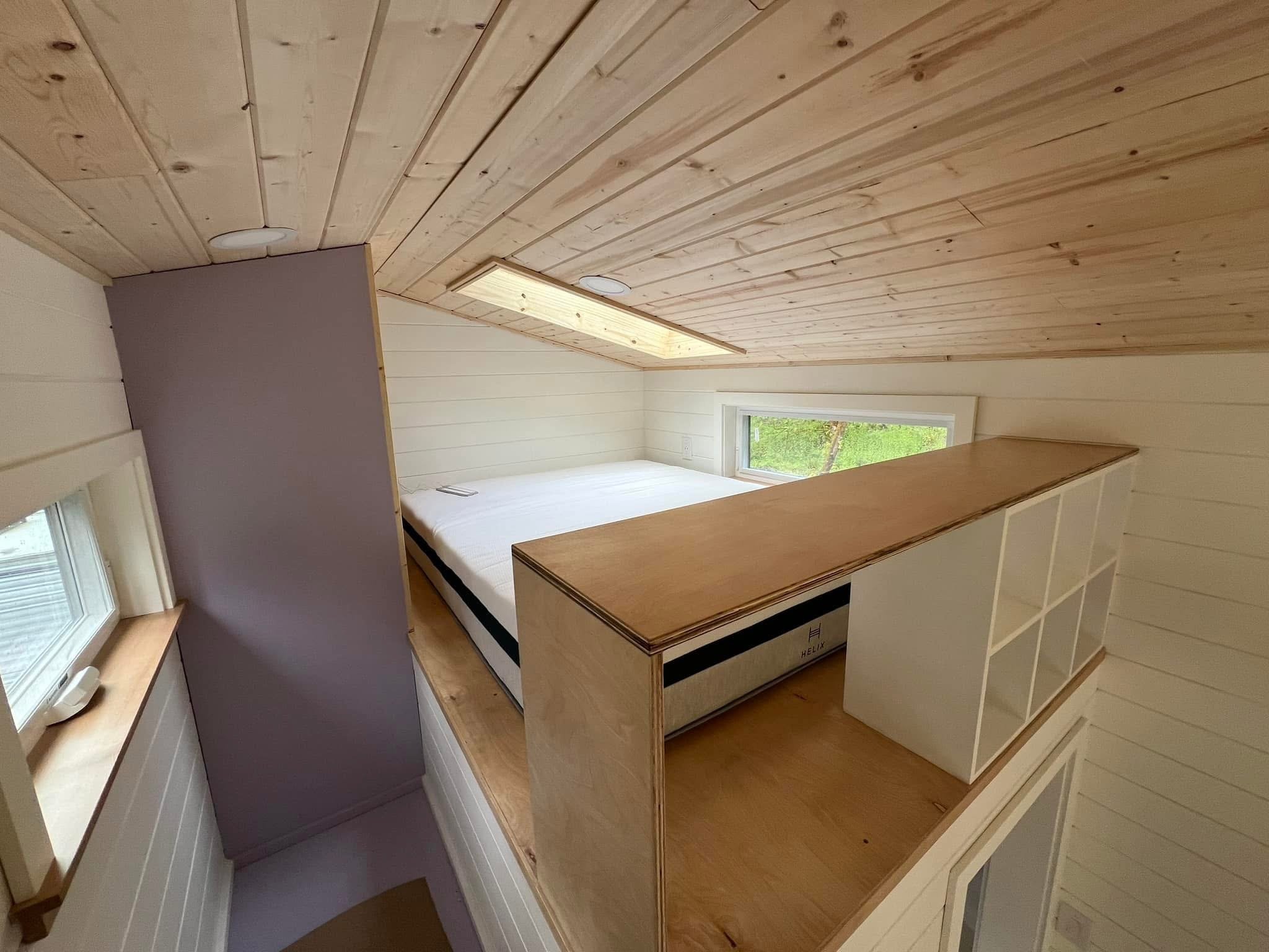 A room with a wooden ceiling, a window showing greenery outside, and a bed frame under construction with a mattress on top.