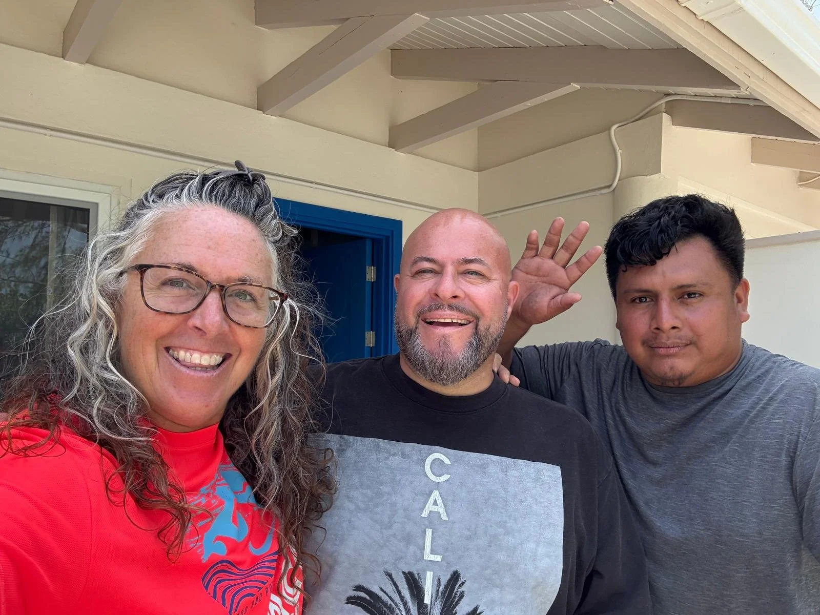 Three people smiling and posing for a selfie outside near a house with a blue door. The woman on the left has curly gray hair, glasses, and is wearing a red shirt. The man in the middle has a beard, is bald, and is wearing a black T-shirt; he is wavi