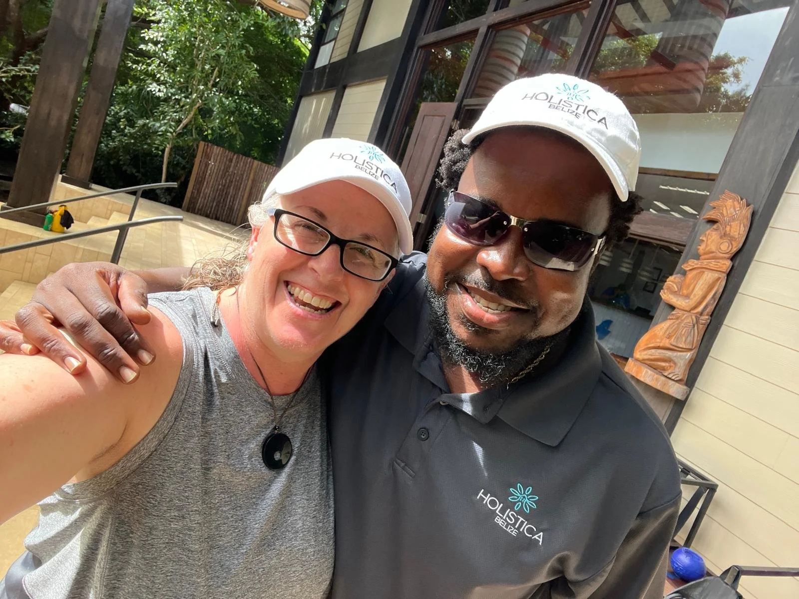 Two smiling people taking a selfie, both wearing white caps with 'HOLISTICA BELIZE' written on them. The person on the left is a woman with glasses and curly gray hair, and the person on the right is a man with dark skin, sunglasses, and a beard. The
