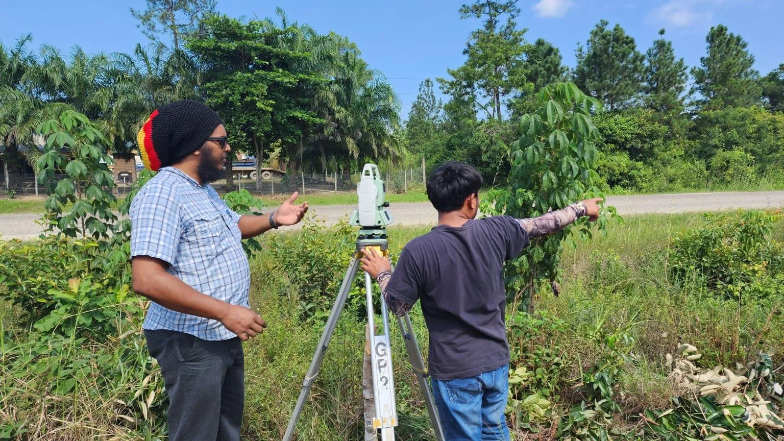 Two men working outdoors, one with a surveying instrument and the other pointing towards the trees.